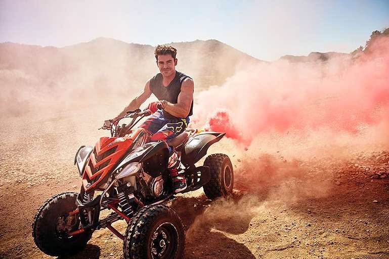 Adult male riding an orange and black all-terrain vehicle (ATV) in a desert landscape with mountains and pink smoke.