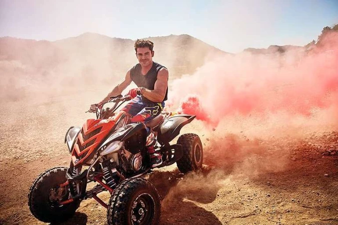 Adult male riding an orange and black all-terrain vehicle (ATV) in a desert landscape with mountains and pink smoke.
