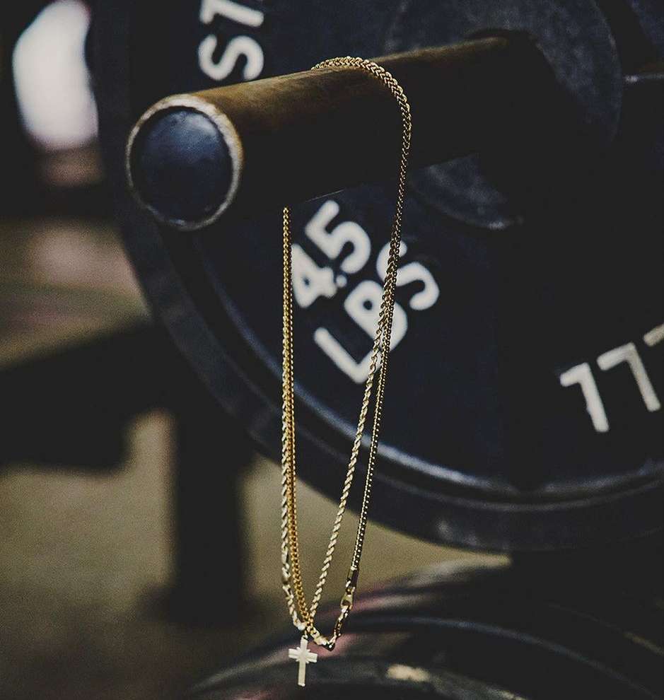 Gold chain necklace with cross pendant hanging on a metal weightlifting bar in a gym, set against a blurred black weight plate.