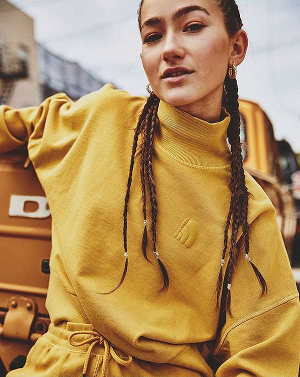 A brunette woman with braids poses in a yellow sweatshirt and matching sweatpants, leaning against a yellow vehicle in an urban setting.
