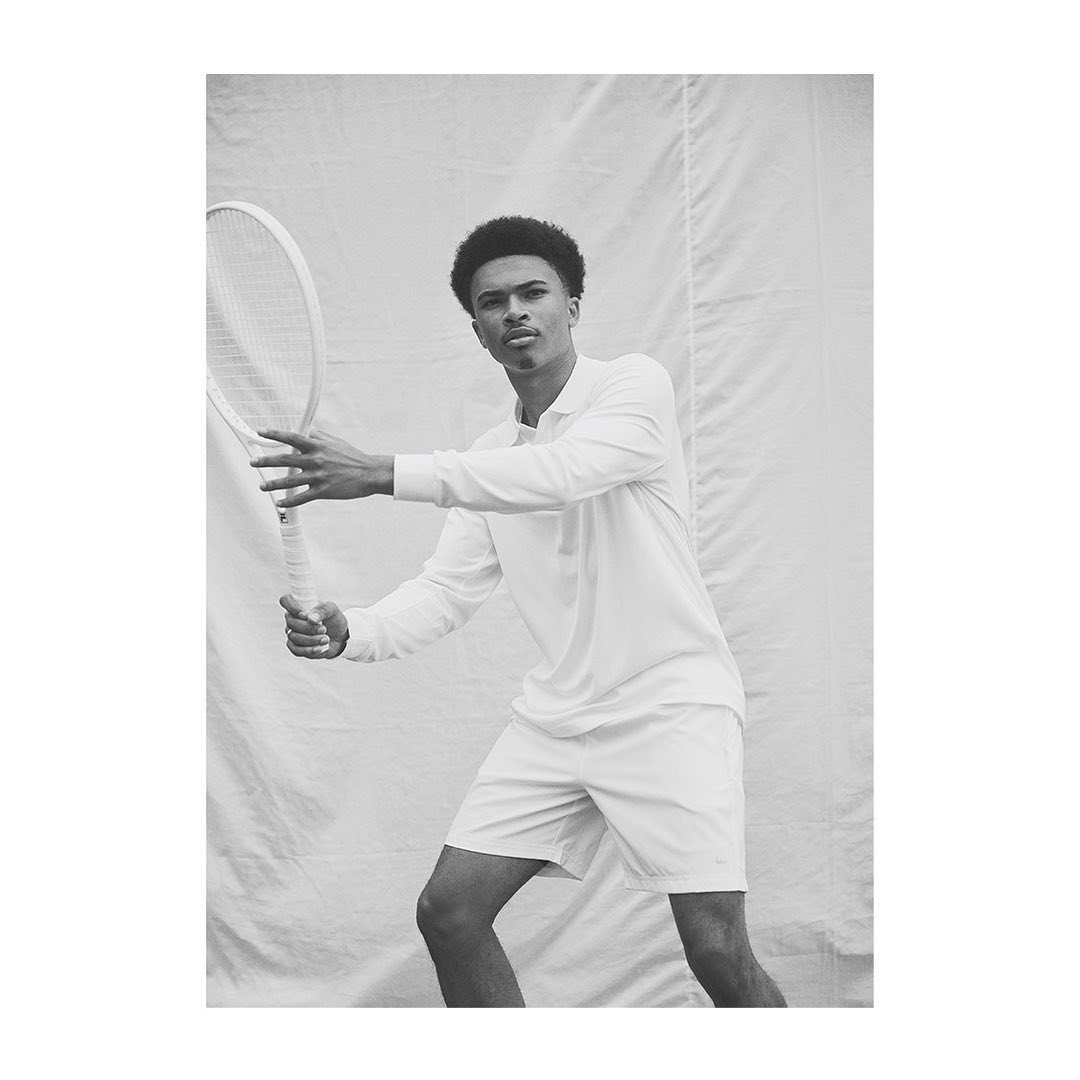 Black and white portrait of a young adult man in a collared shirt and shorts holding a tennis racket against a fabric backdrop.