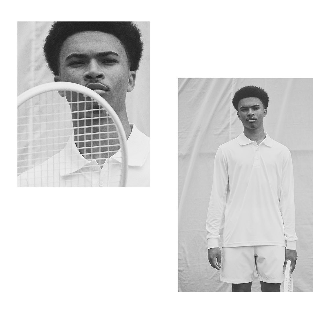 Monochromatic portrait of a young adult man in a white tennis outfit holding a racket against a white draped backdrop in a studio.