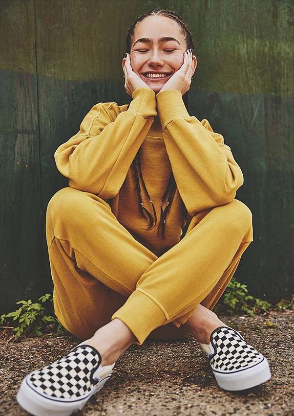 Smiling woman in yellow sweatshirt and pants sits cross-legged against green wall with black-and-white checkerboard Vans slip-on sneakers.