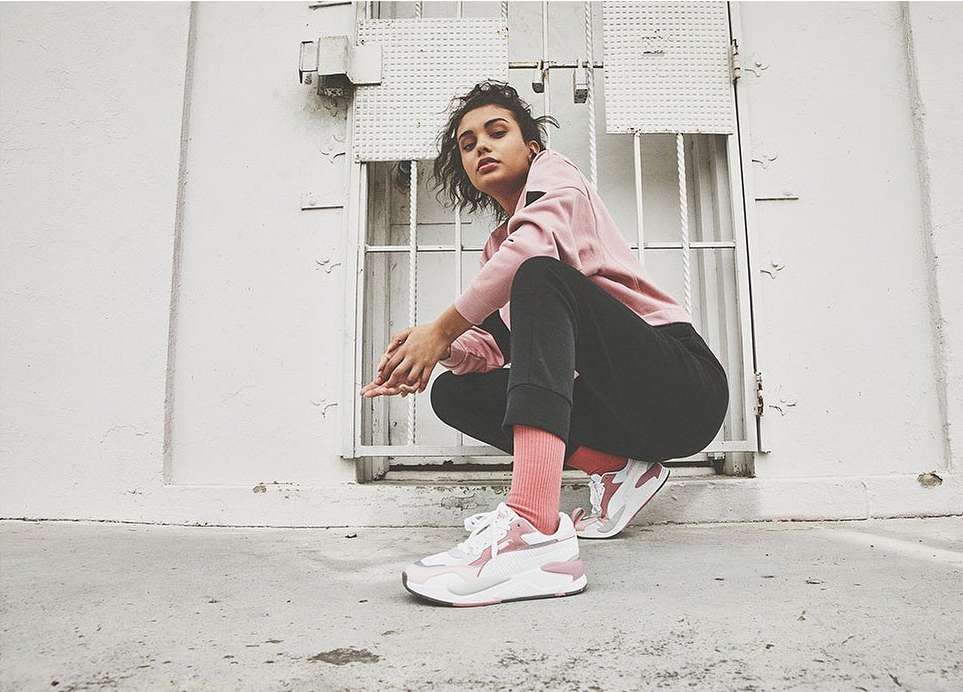 A woman in pink top and black pants crouches, modeling athletic shoes against a white wall with metal grate details outdoors.