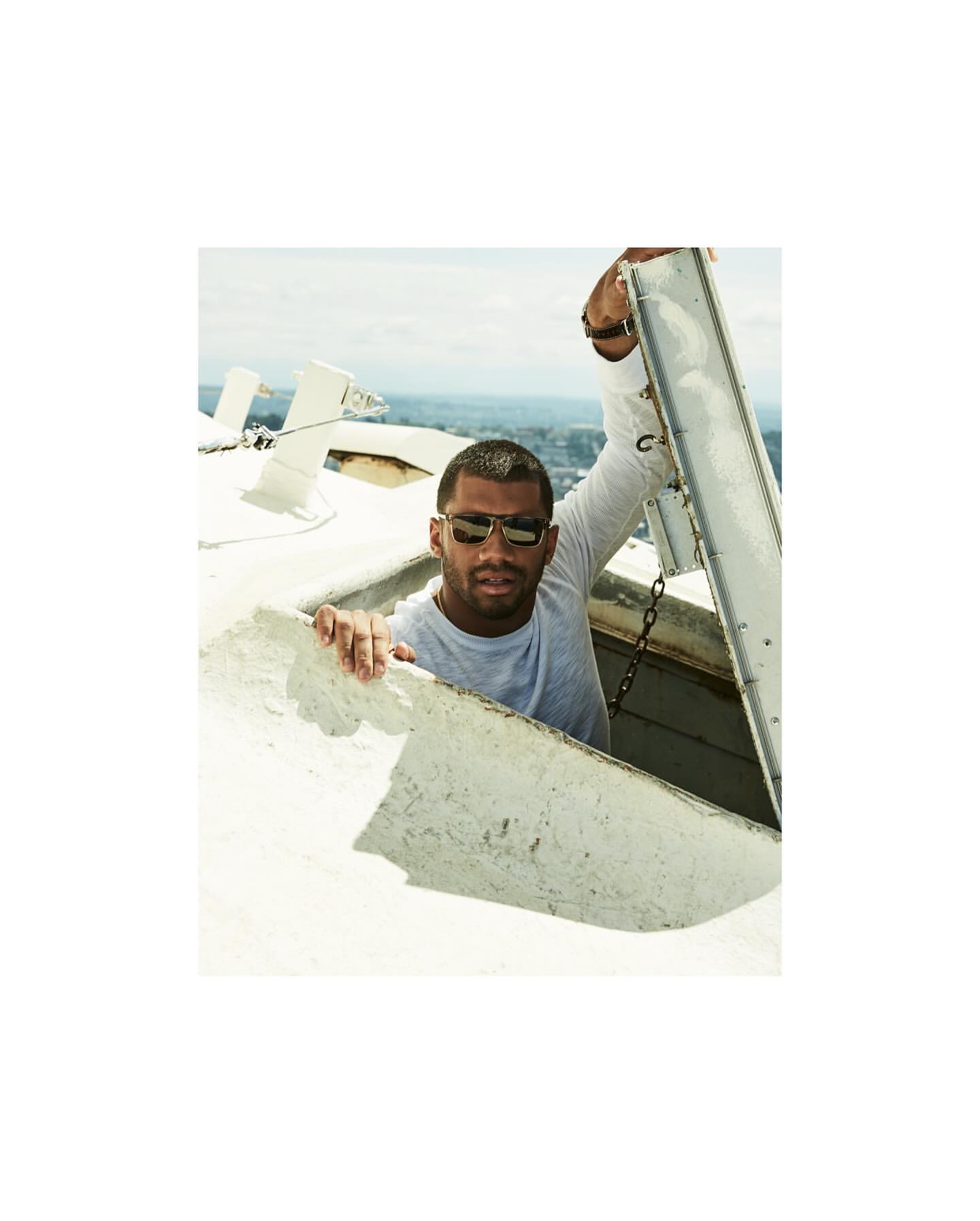 Adult male wearing sunglasses and a long sleeve shirt, looking out of an opening in a tall white structure with an urban background.