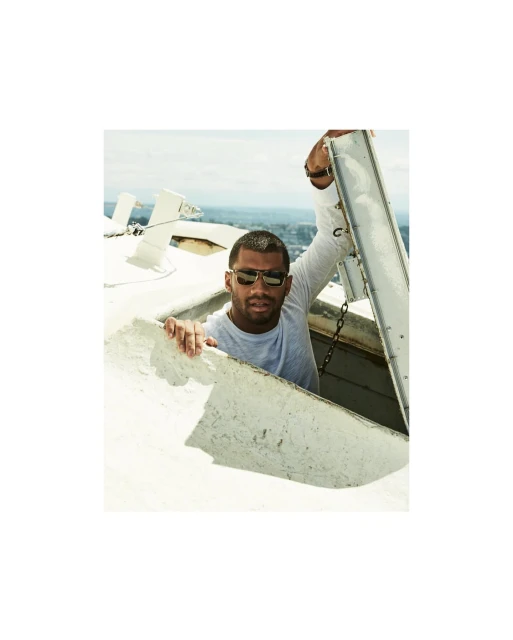 Adult male wearing sunglasses and a long sleeve shirt, looking out of an opening in a tall white structure with an urban background.