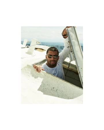 Adult male wearing sunglasses and a long sleeve shirt, looking out of an opening in a tall white structure with an urban background.