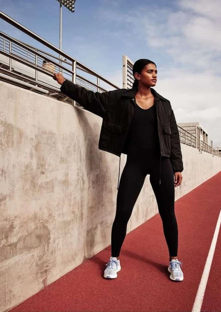 Woman in black athletic wear poses confidently on a red running track with a grey wall and stadium lights against a bright blue sky.