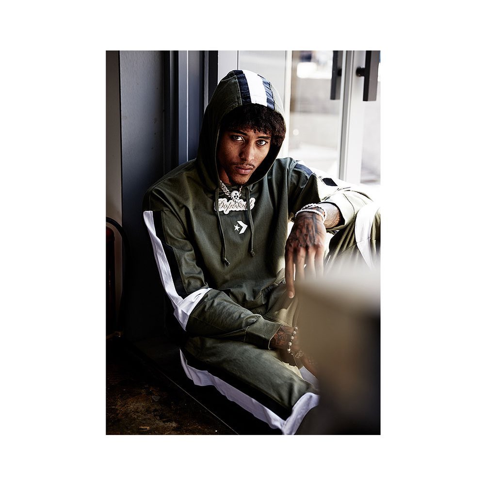 Man in an olive Converse tracksuit, seated and wearing a hooded jacket and silver chain, is lit by natural window light.