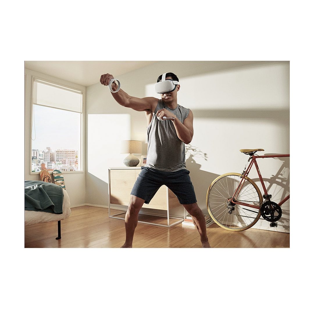 Adult male in tank top and shorts wearing a VR headset with hand controllers, in an indoor setting with a bicycle and furniture.