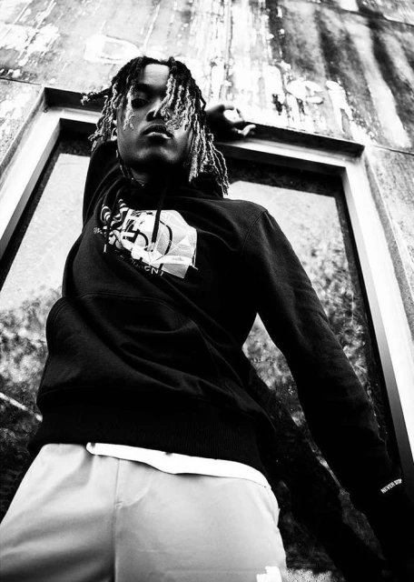 A black and white medium shot of a person with locs wearing a hoodie and light pants while leaning against a textured wall.