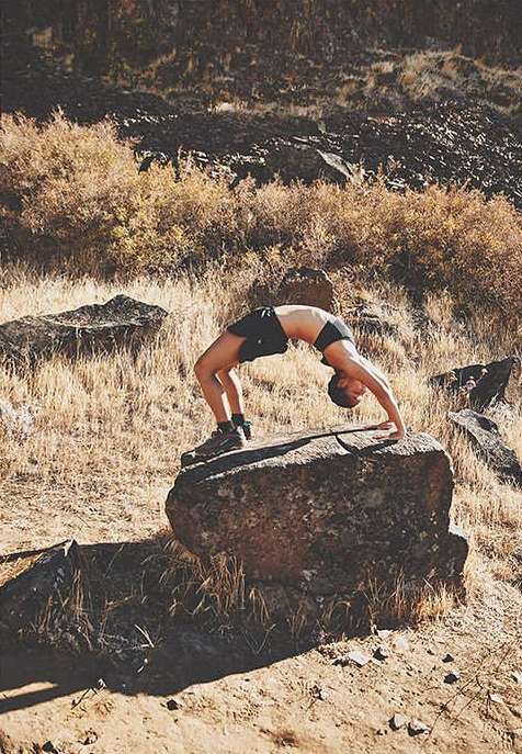 A woman in athletic wear balances in a backbend pose on a large rock in a sunlit outdoor landscape with dry grasses and rocky hills.
