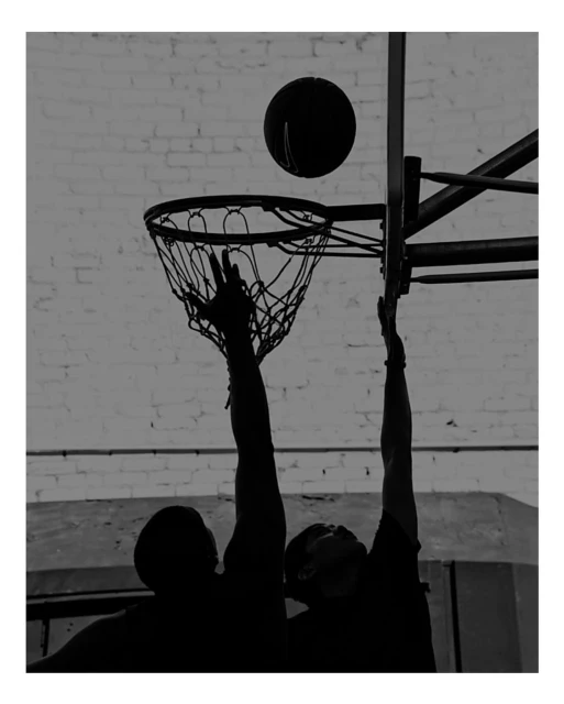 Silhouette of two basketball players reaching for the ball near the hoop against a textured light grey wall in an indoor setting.