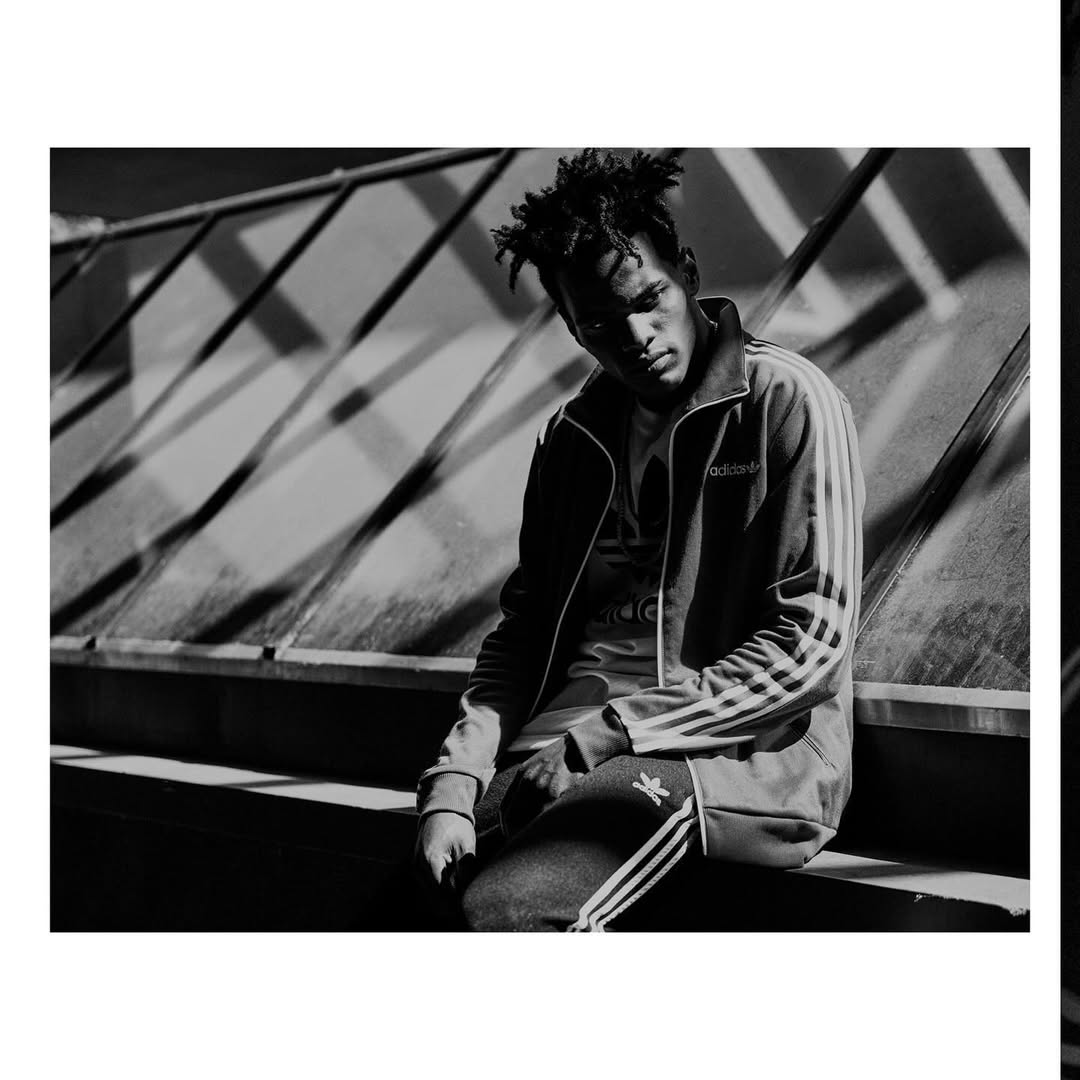 Portrait of a young adult male in black and white, wearing an Adidas tracksuit while seated outdoors with geometric background.