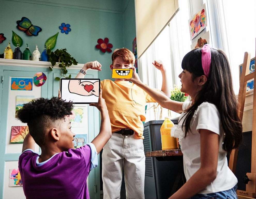 Three children in art studio holding tablets displaying artwork, smiling, surrounded by artwork and art supplies. Indoors, eye-level.