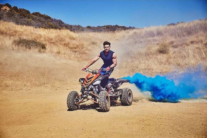 Athletic man rides orange and black quad bike across arid terrain with hills under a clear blue sky. Blue smoke billows behind the ATV.