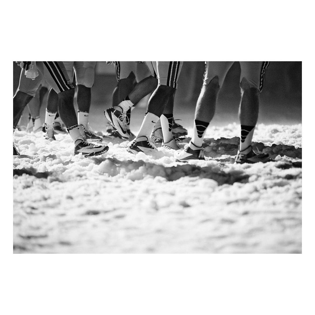 Black and white photo of a line of football players standing on a snowy field with matching socks and sports shoes, eye-level shot.