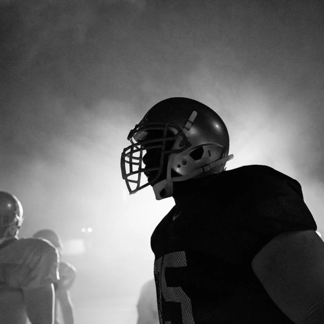 Black and white image of a football player wearing a helmet and shoulder pads, in a smoky environment with other players behind him.