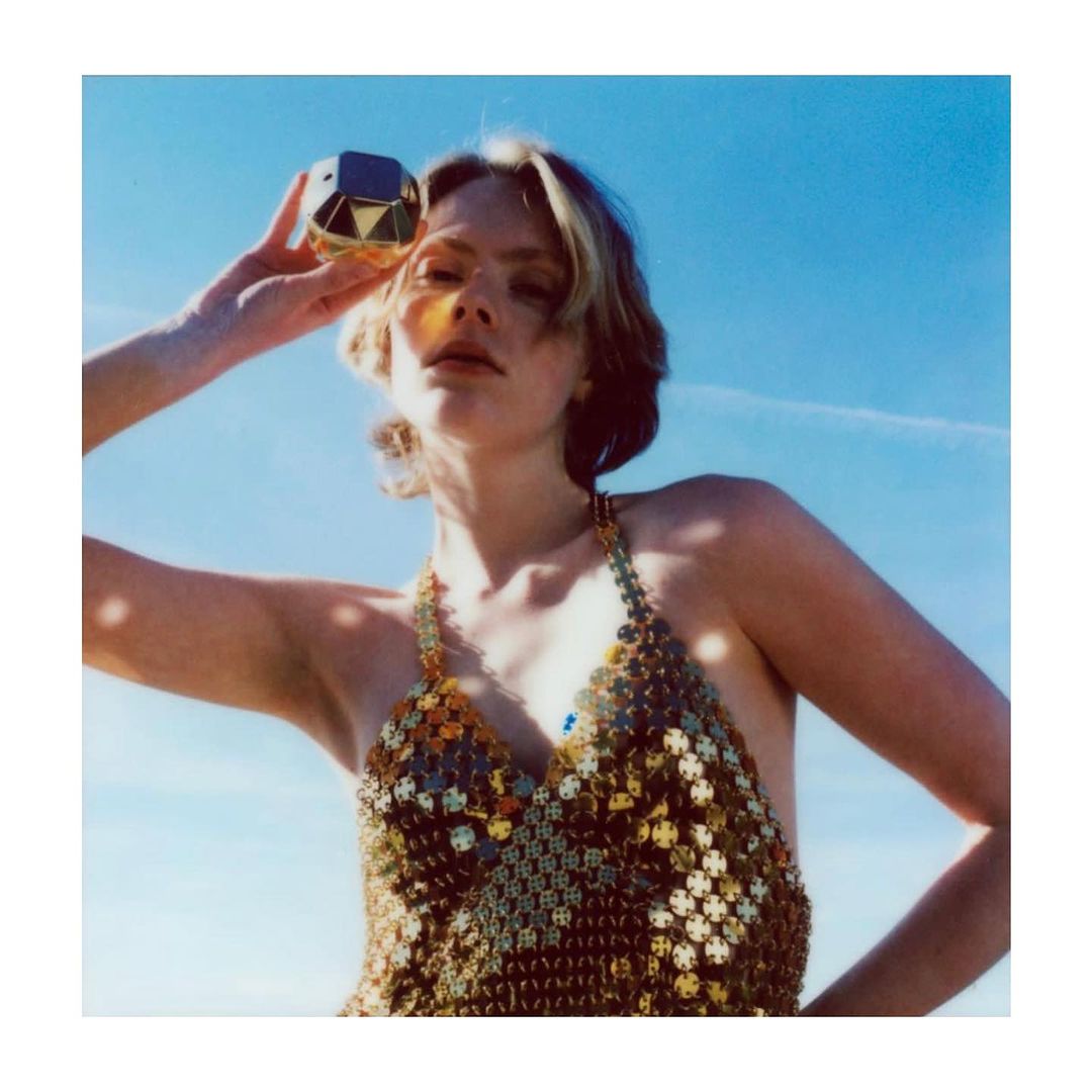 Woman in gold top holds faceted gold object outdoors with blue sky background; beauty, fashion, and product shot.