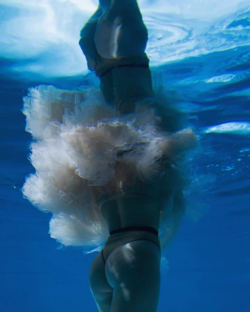 Underwater shot of an adult female in a tan swimsuit and sheer, light-colored skirt. The person is submerged in clear blue water.
