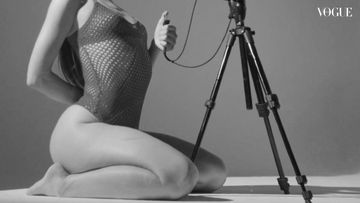 Woman in swimsuit kneeling next to a camera on a tripod in a studio with natural lighting and the Vogue logo visible.