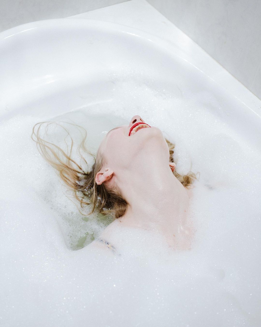 Woman relaxes in a bubble bath with her head tilted back. The white bathtub is filled with water and fluffy white bubbles.