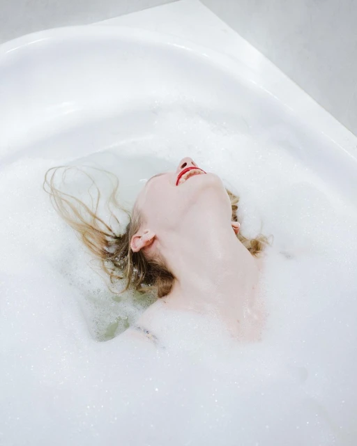 Woman relaxes in a bubble bath with her head tilted back. The white bathtub is filled with water and fluffy white bubbles.