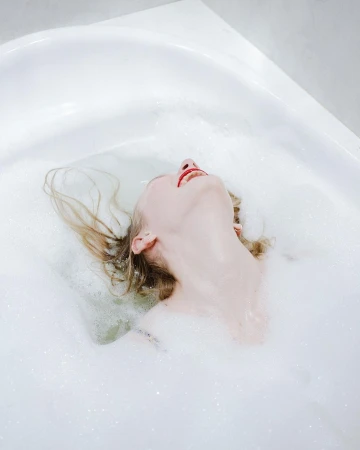 Woman relaxes in a bubble bath with her head tilted back. The white bathtub is filled with water and fluffy white bubbles.