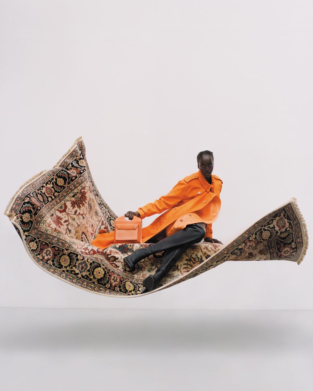 Woman in orange jacket sits on an ornate flying carpet, holding a peach-colored purse, against a clean white backdrop.
