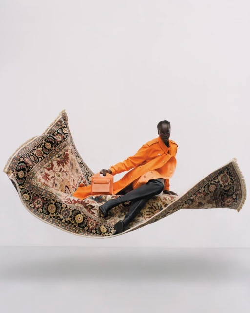 Woman in orange jacket sits on an ornate flying carpet, holding a peach-colored purse, against a clean white backdrop.