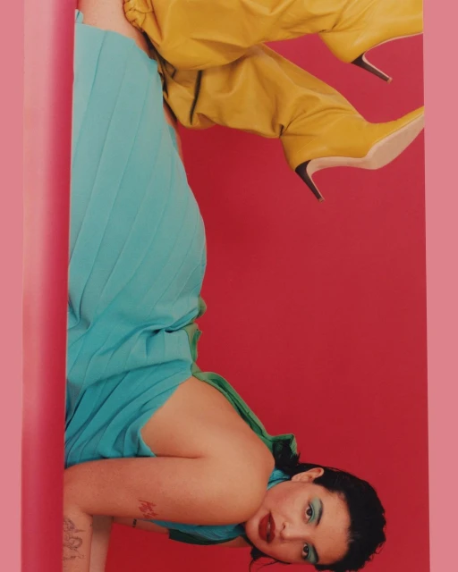 Portrait of a woman in a turquoise dress and yellow boots posing against a pink and red background in a studio setting.