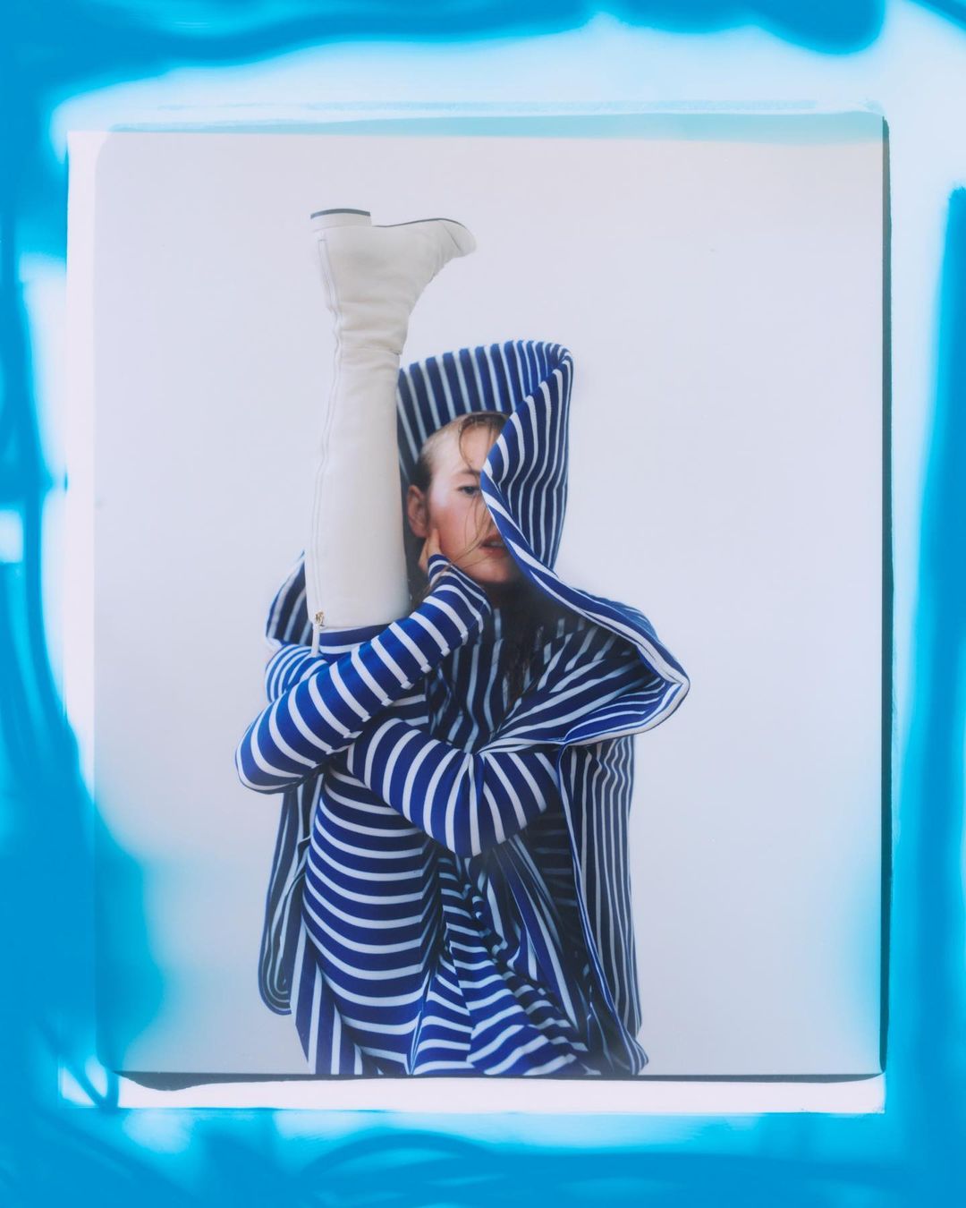 Woman in striped hooded jacket and white boots poses creatively in a brightly lit indoor setting with an abstract frame.