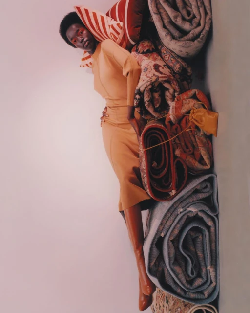 A fashion-forward photograph presents a Black woman in a yellow dress, reclining against a stack of rolled carpets in a brightly lit studio.