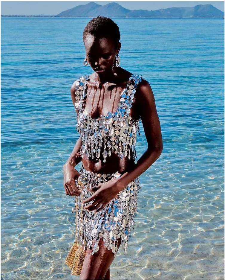 A dark-skinned female model in a silver sequin two-piece stands in shallow ocean water against a bright, sunny backdrop for a fashion editorial.