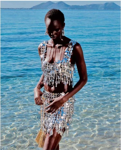 A dark-skinned female model in a silver sequin two-piece stands in shallow ocean water against a bright, sunny backdrop for a fashion editorial.