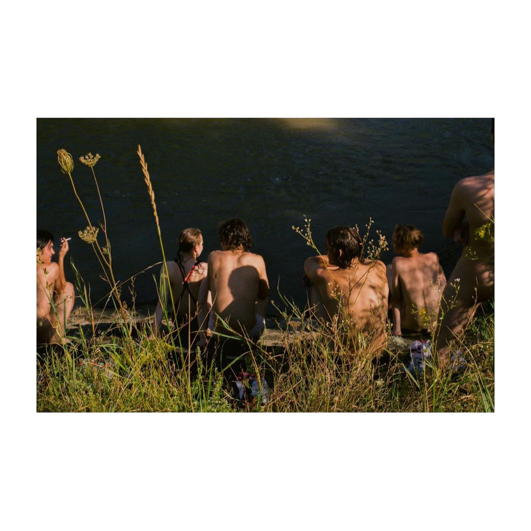 Group of six people sit on grassy bank by a dark river, backs to camera, bathed in sunlight, creating an atmospheric outdoor scene.