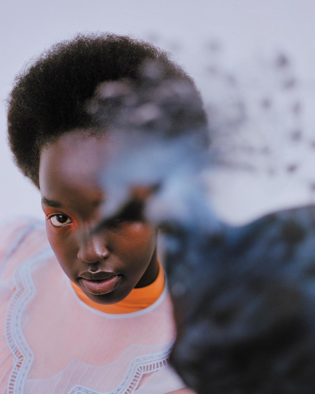 Striking studio portrait of a woman with dark skin and afro, wearing a sheer, pale-pink top. Dominant blue, white backdrop.
