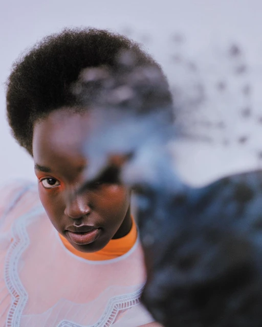 Striking studio portrait of a woman with dark skin and afro, wearing a sheer, pale-pink top. Dominant blue, white backdrop.