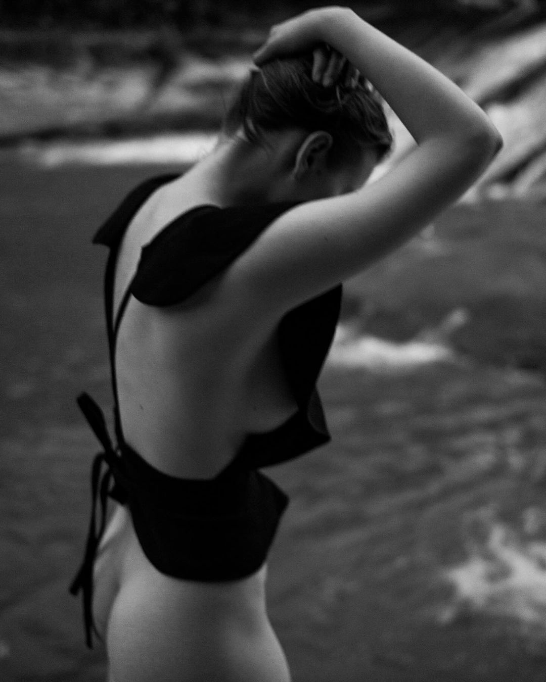 Adult female with her hand on her head poses wearing a black garment, standing outdoors in front of a blurred river background.