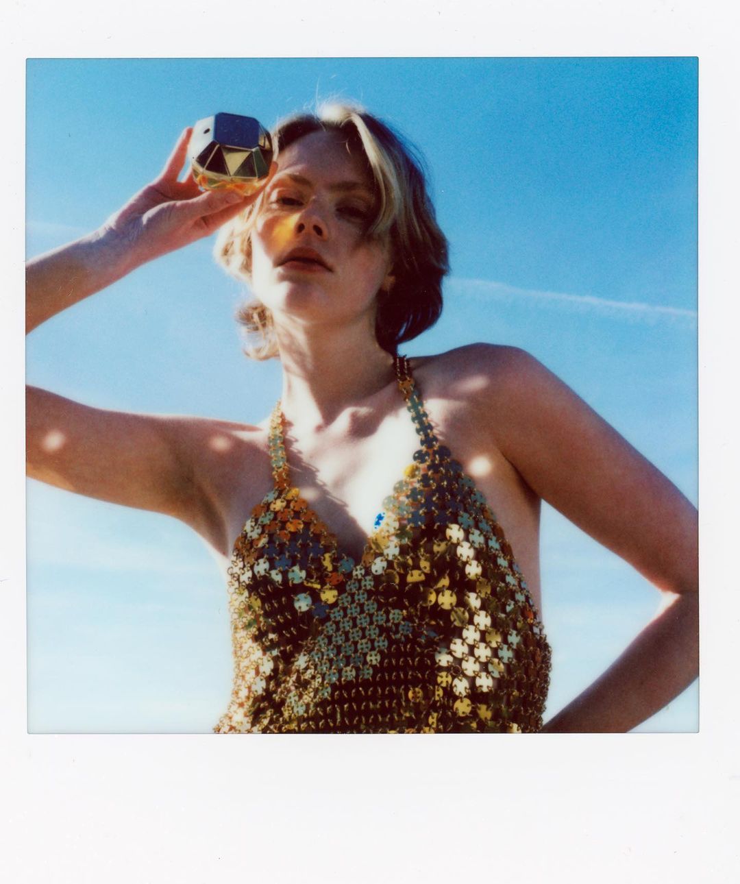 Young woman poses outdoors in a chainmail top, holding a gold perfume bottle to her forehead against a clear blue sky backdrop.