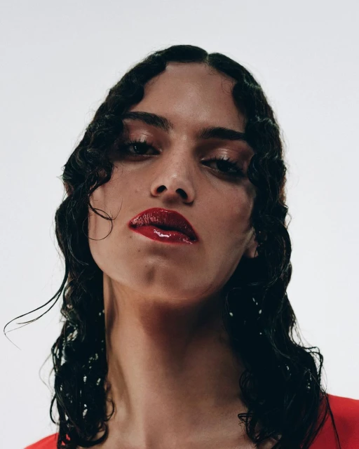 Close-up portrait of a young adult woman with wet, styled dark hair, glossy red lipstick, and minimal eye makeup against a plain white background.