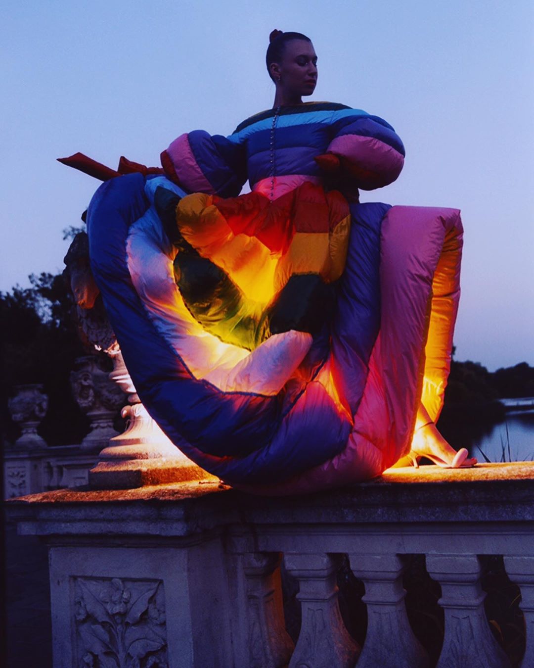 Fashionable adult female poses in a whimsical rainbow gown, outdoors at dusk by a stone wall with trees and water in the distance.