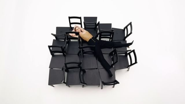 Woman poses on a structure of many black chairs in a brightly lit studio. She wears dark pants and a light-colored top. High-angle shot.