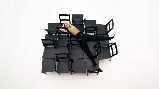 Woman poses on a structure of many black chairs in a brightly lit studio. She wears dark pants and a light-colored top. High-angle shot.