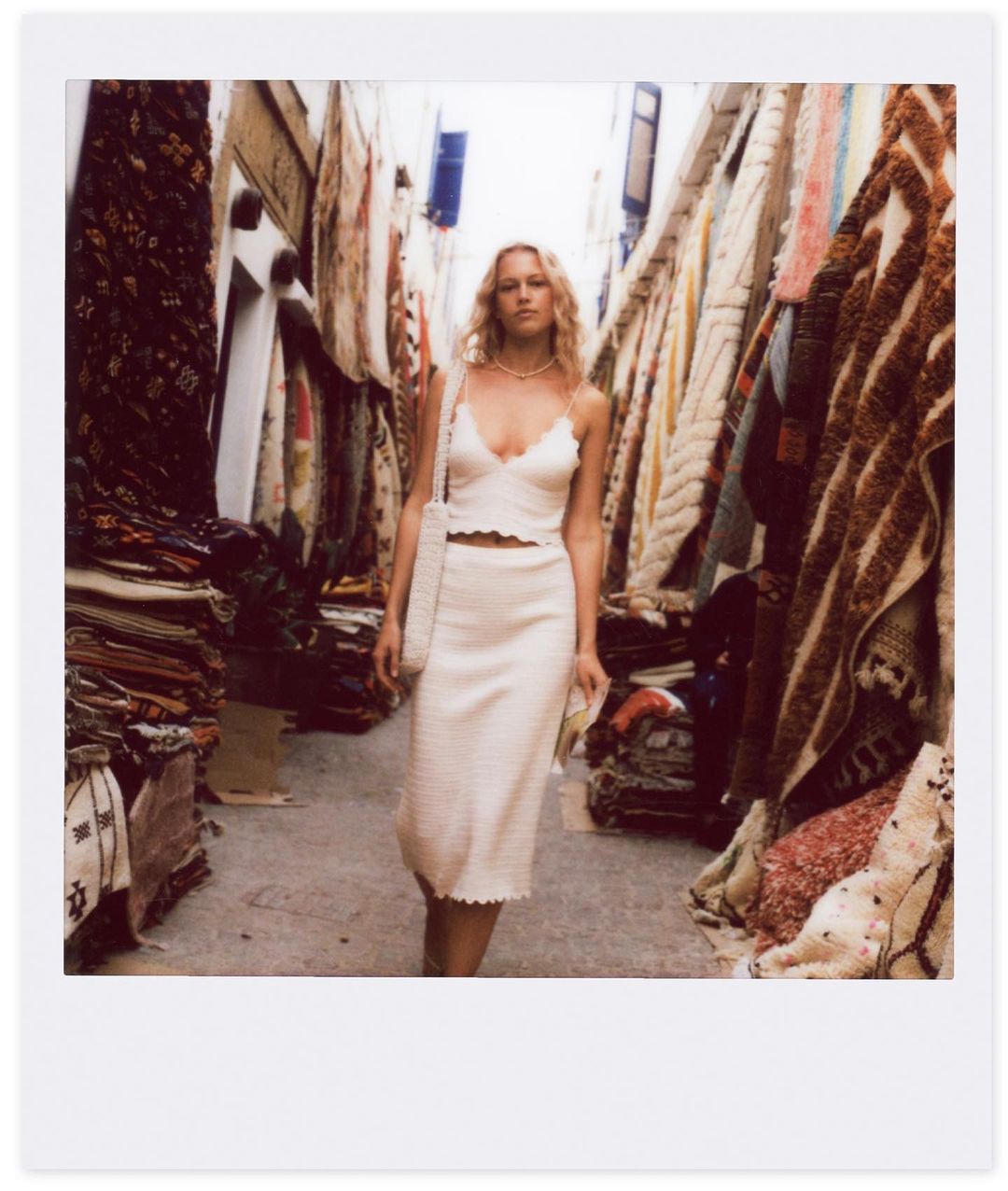 A blonde woman in a cream knit skirt and top walks down a narrow alley lined with stacked carpets in the daytime.
