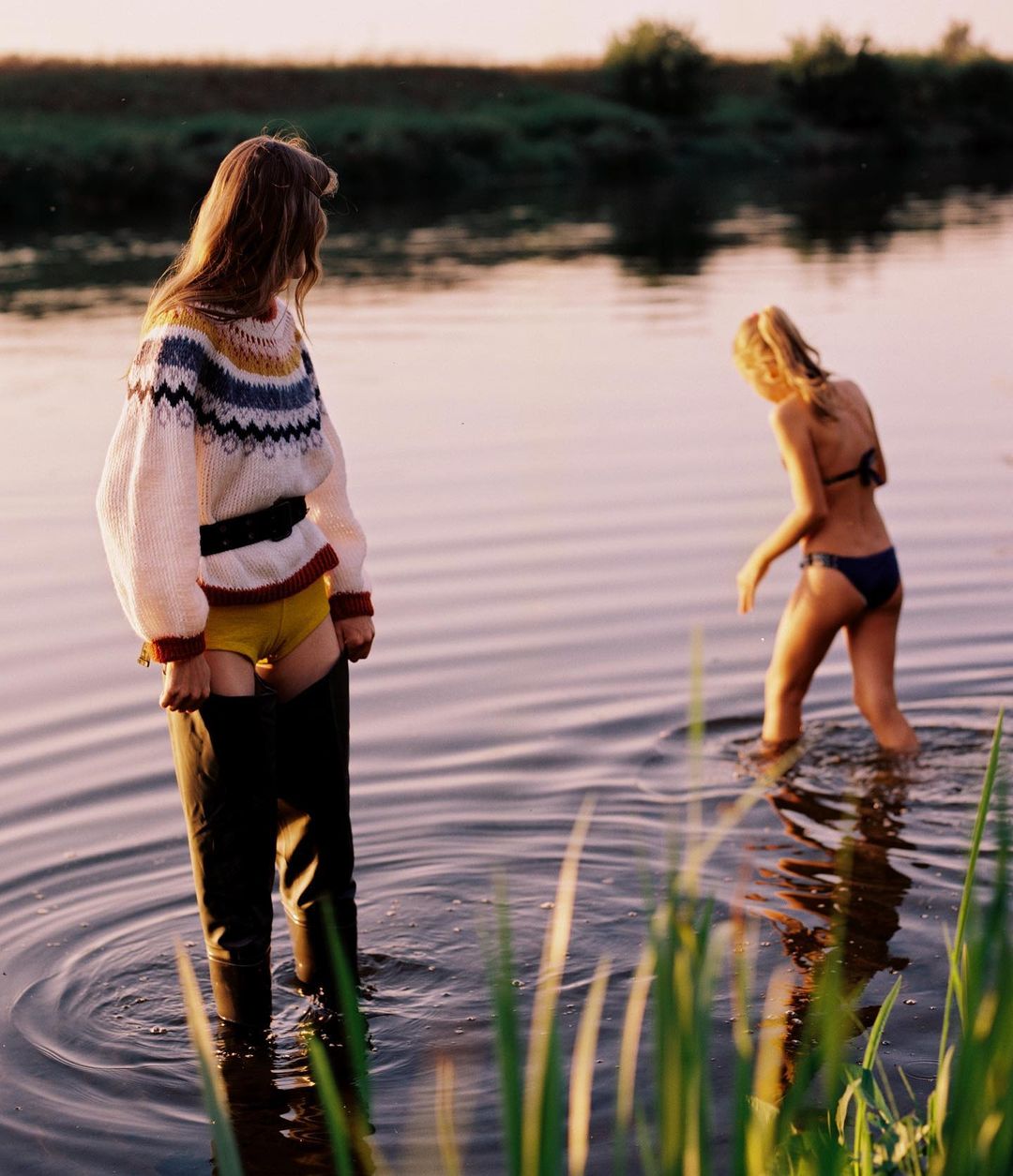Two young women wade in a lake at dusk. One wears a patterned sweater and waders, and the other wears a bikini, creating a summery outdoor scene.