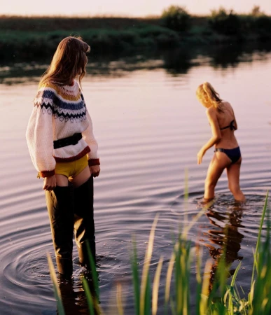 Two young women wade in a lake at dusk. One wears a patterned sweater and waders, and the other wears a bikini, creating a summery outdoor scene.