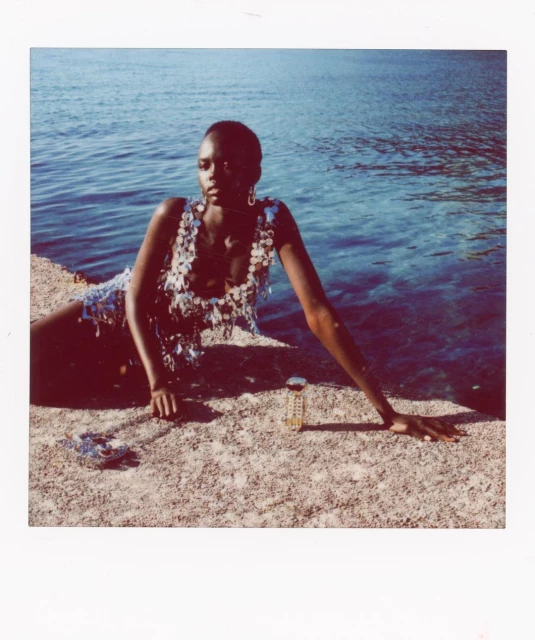 Woman in a sequined top poses on a concrete surface by the ocean, wearing hoop earrings and with a small bottle beside her.