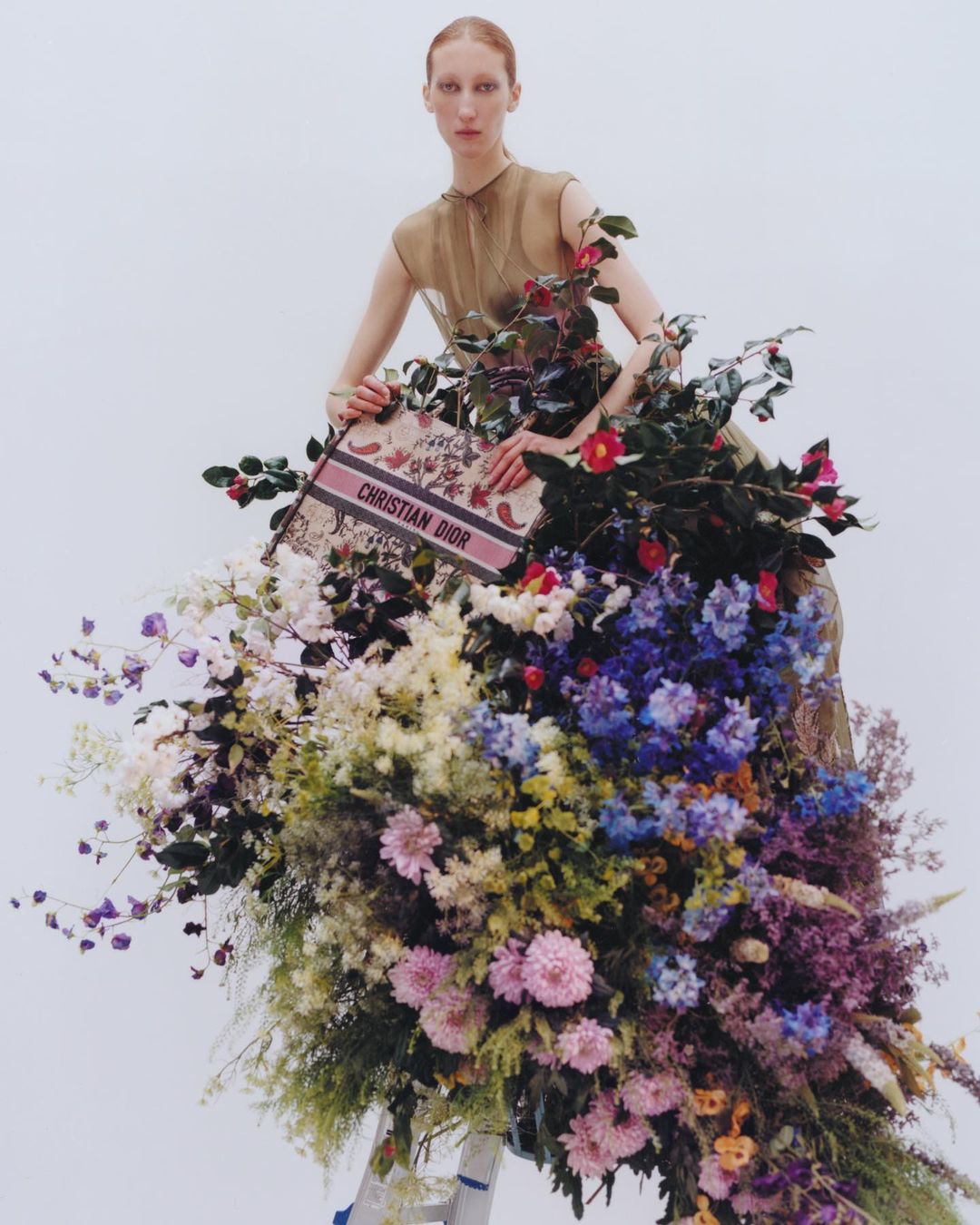 Fashion editorial shot of a tall, slender woman in an airy dress posing with a floral arrangement and a Christian Dior branded bag.
