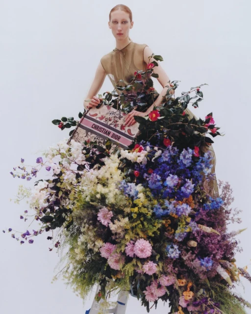 Fashion editorial shot of a tall, slender woman in an airy dress posing with a floral arrangement and a Christian Dior branded bag.
